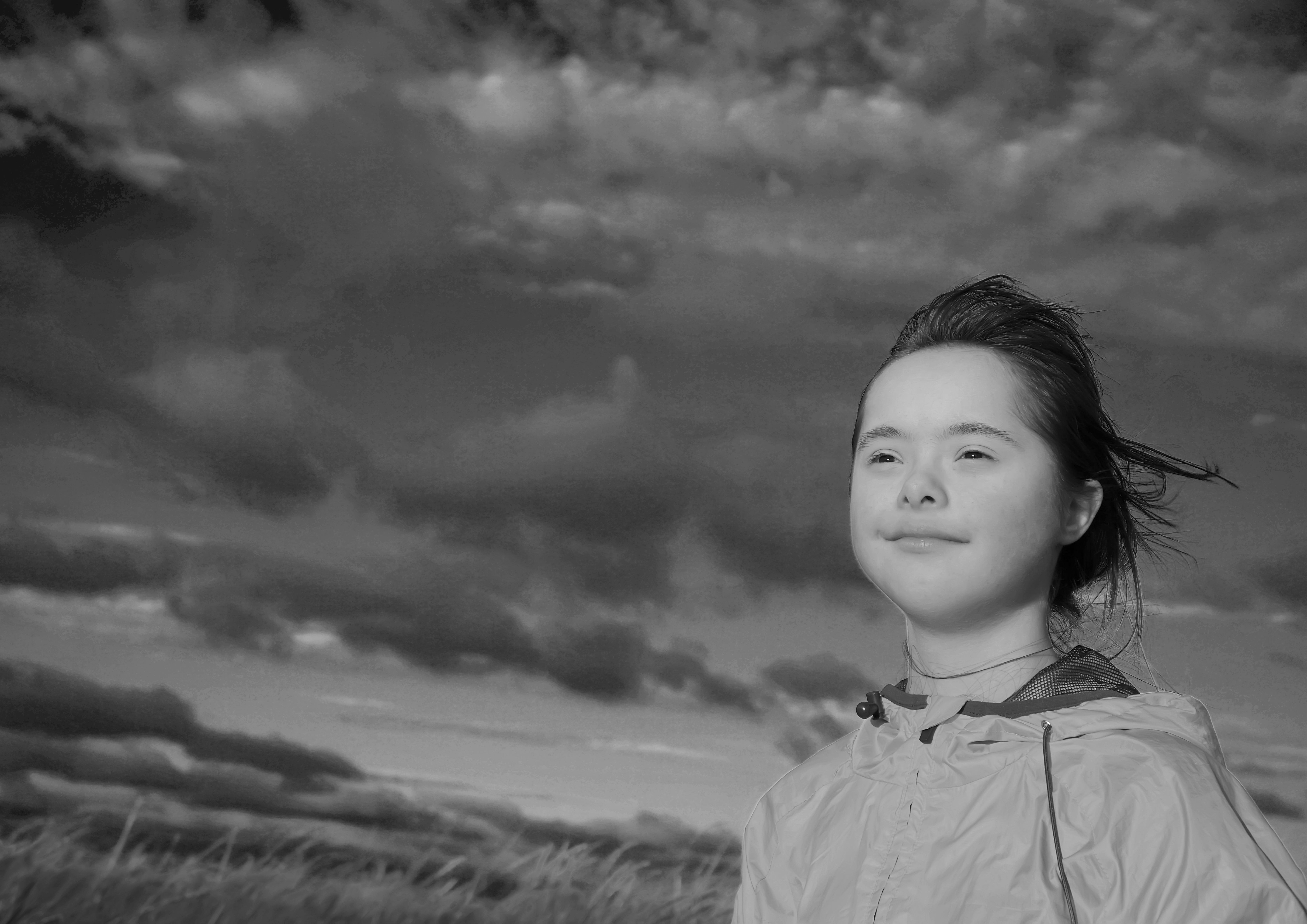 Looking slightly up at a young person who is standing in a field and looking upwards with a content look on their face. A dramatic dark cloudy sky fills most of the frame.
