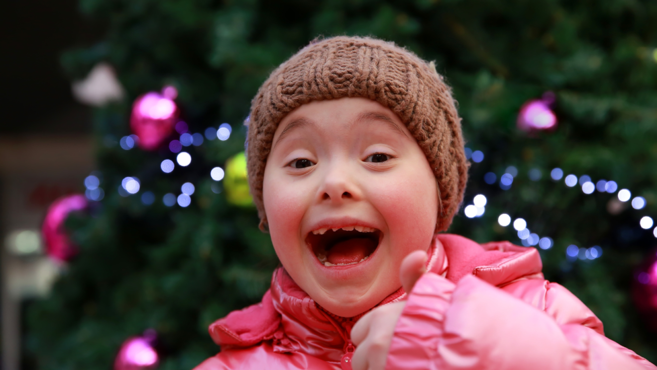 A young girl with Down Syndrome smiles widely at the camera and holds her thumb up. She is wearing a beanie and a bright pink jacket.