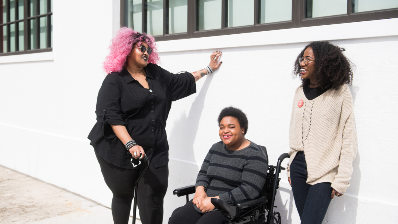 Three Black and disabled folx (a non-binary person resting one hand on the wall and the other on her cane, a non-binary person sitting in a power wheelchair, and a femme leaning while standing) rest and chat outdoors on a sunny day.