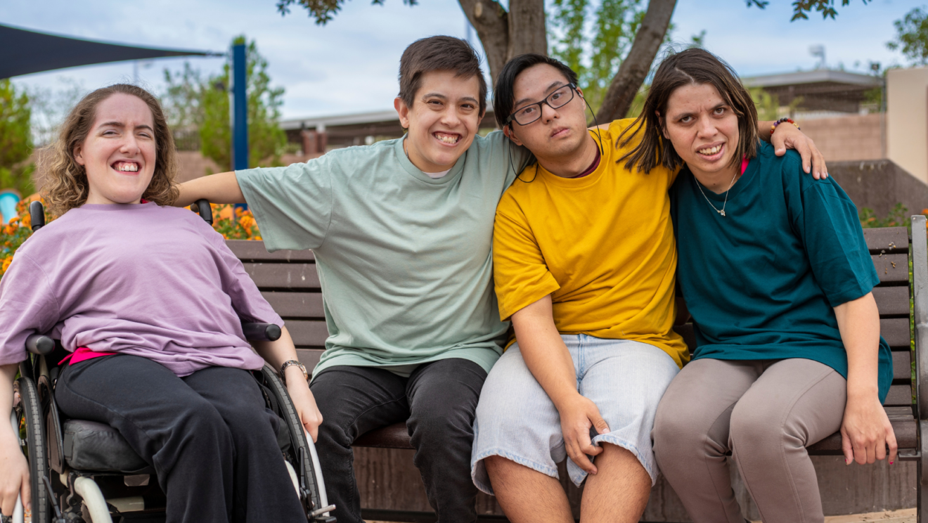 Four young people with disability smiling in a park with their arms around each other.