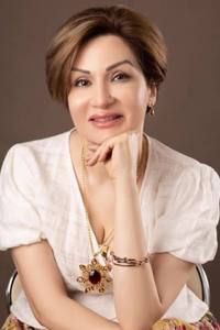 Zhila sitting with her chin resting on her hand and smiling slightly. She is wearing a long gold necklace with a large pendant and a white shirt.