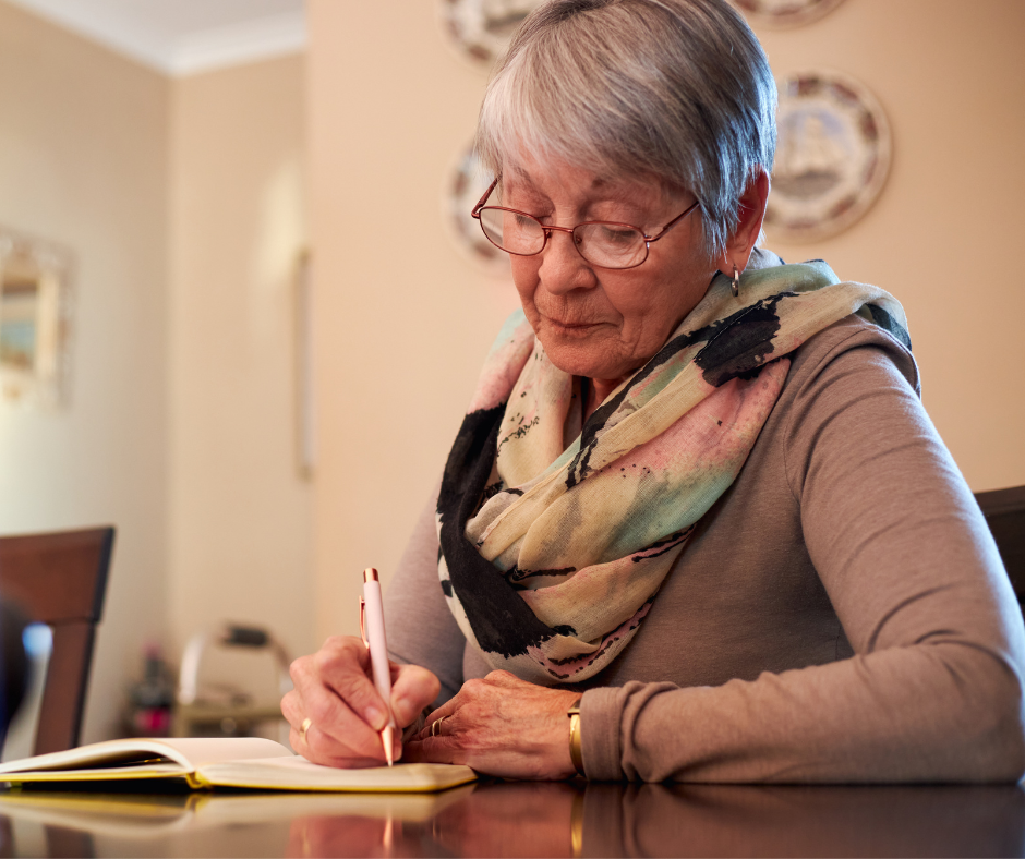An older woman writing in a book at a dining table. She is wearing glasses and a colourful scarf.