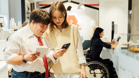 A man and woman look at a mobile phone screen and in the background a woman using a wheelchair looks at an electronic hand device