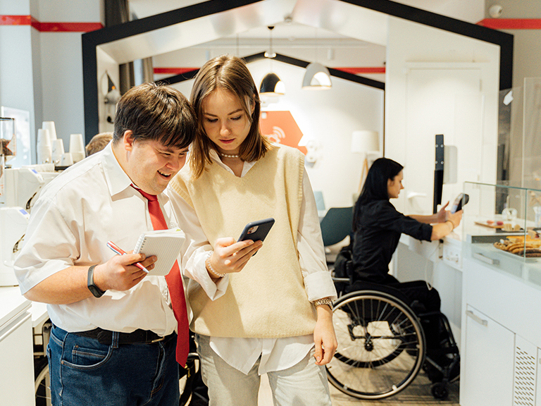 [IMAGE a man and woman look at a mobile phone screen and in the background a woman using a wheelchair looks at an electronic hand device]