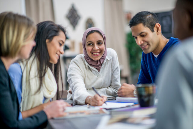 A multi-ethnic group of adults are indoors in a design studio. They are sitting around a table and making business plans. A Muslim woman wearing a headscarf is writing her ideas in front of the group.