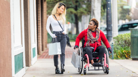 A young black disabled woman with a wheelchair and a bright colored sweater and her Asian friend walk around the city.