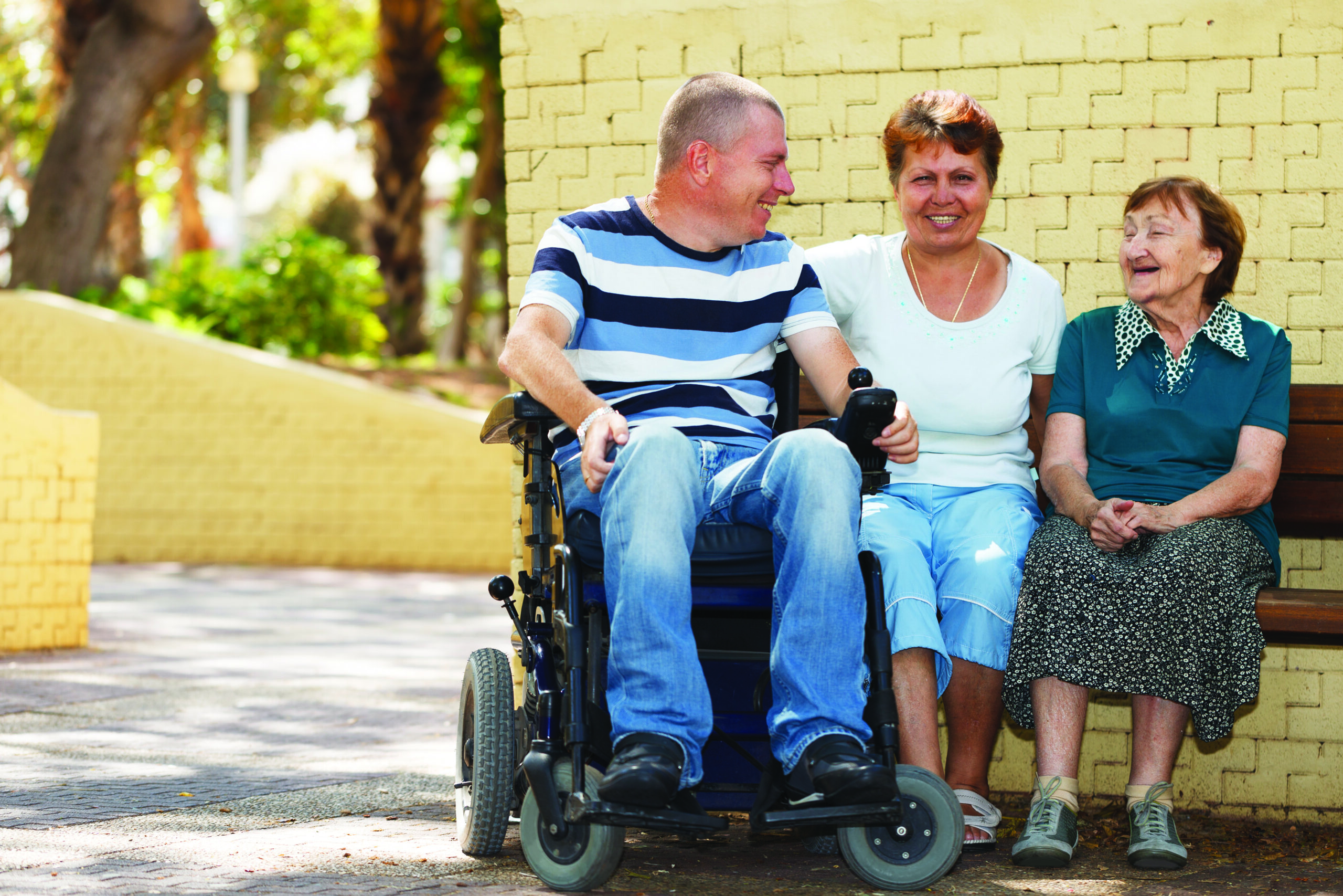 three people smiling at each other. one is sitting in a wheelchair and the other two are seated on a bench