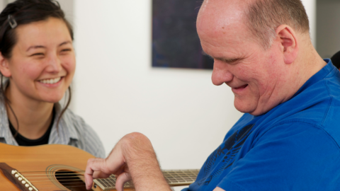 A man plays a guitar while a smiling woman watches him