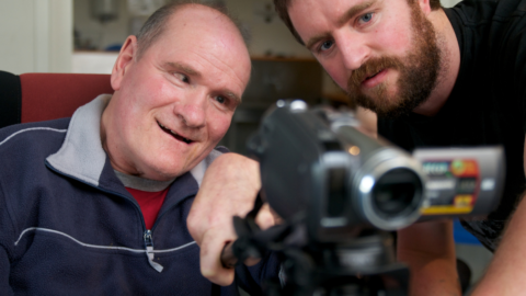 Man assisting another man with disability to operate a video camera.