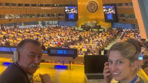 Lisa is sitting in the UN General Assembly with Grant Lindsay, the President and Chair of the Australian Federation of Disability Organisations. Grant is wearing a black jacket and has an ear piece on his ear. Lisa is wearing a green dress and is holding an ear piece to her ear.