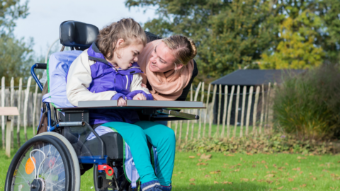 A girl leans over another young girl in a wheelchair.