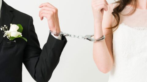 A bride and groom in wedding attire are handcuffed together at the wrist.