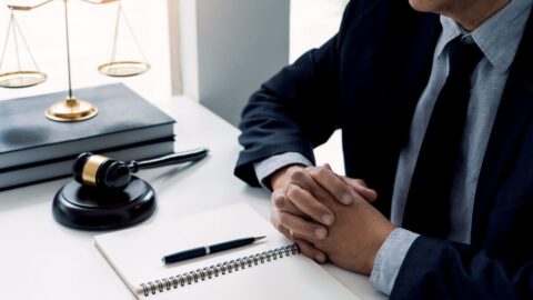 Image shows a person's hands folded on a desk with a gavel and scales of justice next to them.