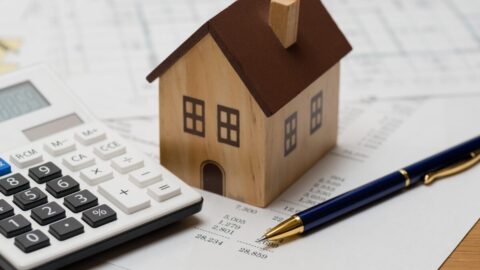 Image shows a small wooden house on top of a pile of bills with a calculator