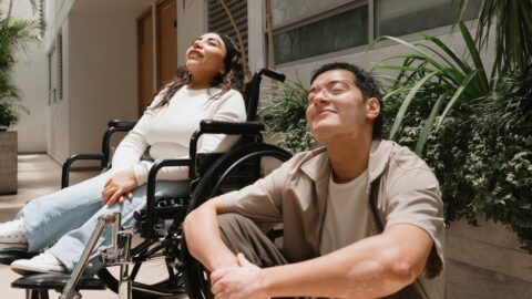 Two people are outside of accommodation enjoying the sunshine. One of them is a wheelchair user and the other is seated on the ground.