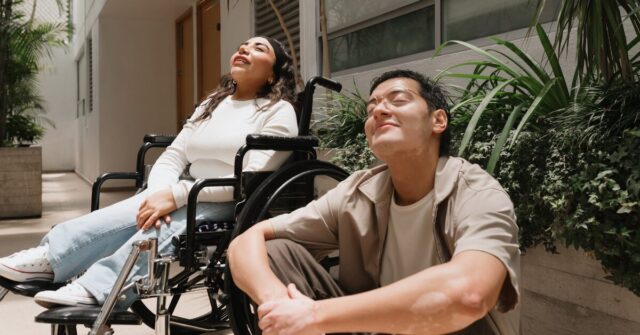 Two people are outside of accommodation enjoying the sunshine. One of them is a wheelchair user and the other is seated on the ground.