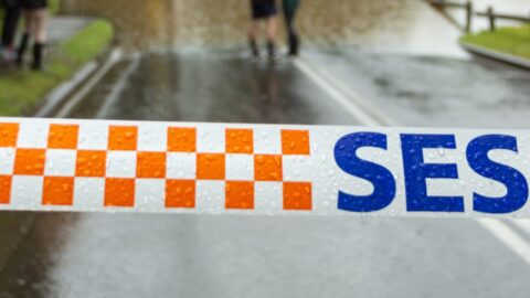 People with umbrellas and gumboots stand on the edge of a road that is closed due to flood with SES tape in the foreground