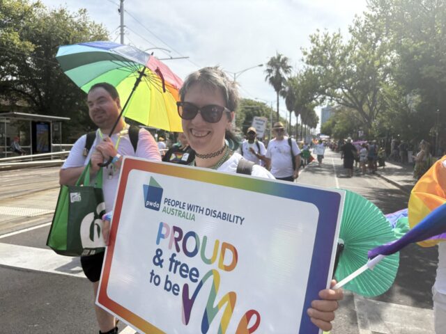 Marchers at Midsumma Pride Festival holding rainbow umbrella and a sign that says Proud & free to be me. 