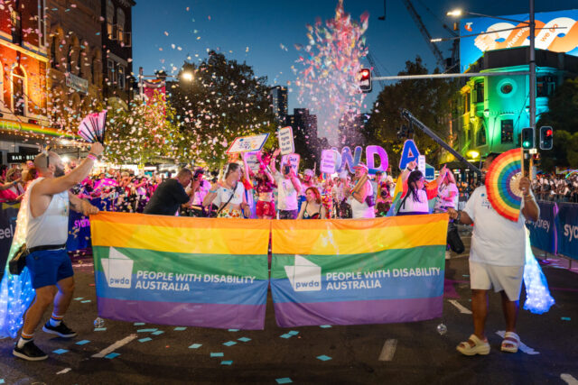 PWDA members and staff cheer behind PWDA branded rainbow flags as confetti cannons go off around them.