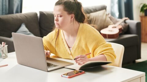 Woman sitting at a laptop wearng a yellow jumper. She has a notebook open and a pen in hand