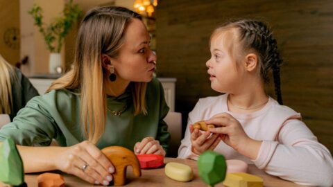 A woman and a young girl are sitting at a table with wooden toys scattered on it. They are both holding a toy each as they look each other in the eyes.