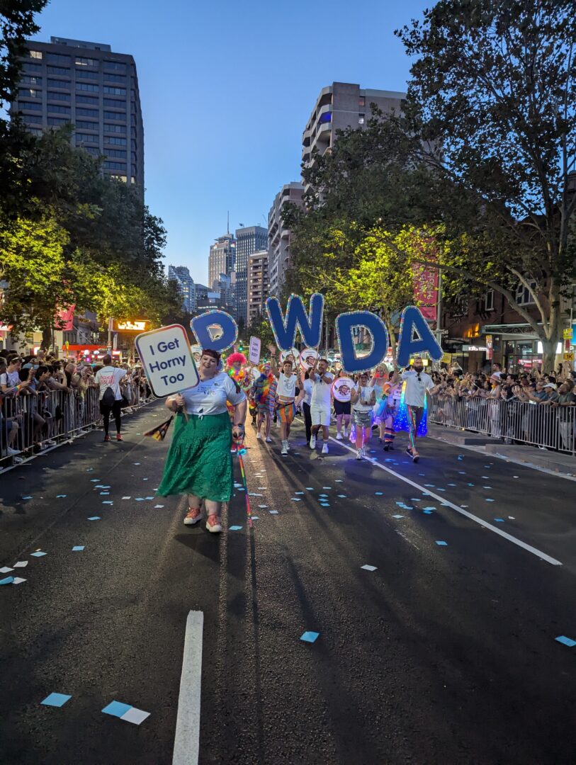 Image shows PWDA members walking in Sydney Gay and Lesbian Mardi Gras parade. People are holding large letters that spell PWDA and a sign that reads I Get Horny Too.