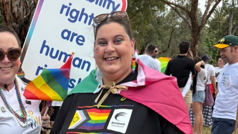 Photo of Shannon Meilak at the 2025 Midsumma Pride March. Shannon is wearing a black pride t-shirt, rainbow cape and is holding on to a black stroller where Shannon’s little black dog is sitting. People are in the background holding PWDA signs and wearing rainbow capes.