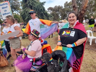 Shannon Meilak_2 - People with Disability Australia Photo of Shannon Meilak at the 2025 Midsumma Pride March. Shannon is wearing a black pride t-shirt, rainbow cape and is holding on to a black stroller where Shannon’s little black dog is sitting. People are in the background holding PWDA signs and wearing rainbow capes.