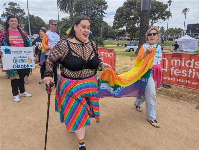 Image shows a group PWDA members participating in the Midsumma march. The group is being led by two people carrying a rainbow PWDA flag.