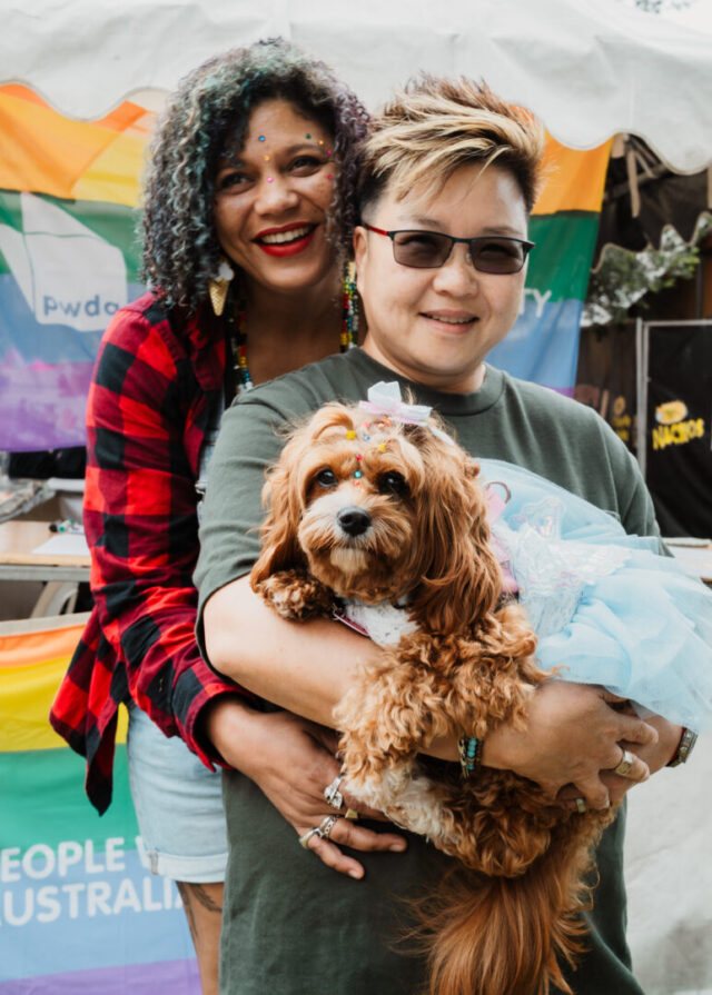 Two people smiling for a photo at Fair Day. They are standing in front of a rainbow PWDA banner. One person stands behind the other with their arms around them, while the person in front holds a small curly brown dog wearing a decorative tutu and hair bow.
