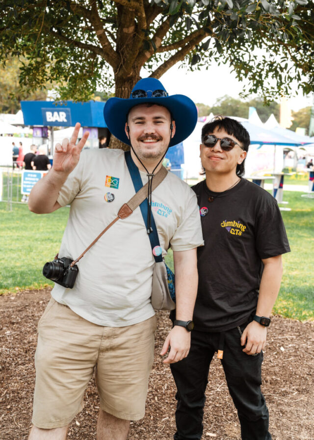 Two people smiling at Fair Day. They are standing under a tree. One wears a bright blue cowboy hat and holds their fingers up a peace sign, with a camera around their neck, while the other wears sunglasses and a black T-shirt.