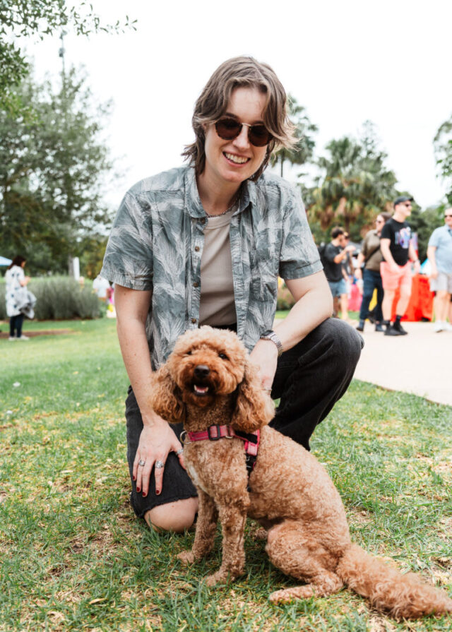A person crouching on the grass at Fair Day, they are wearing sunglasses, smiling and holding a small curly brown dog wearing a pink harness. Trees and other festival attendees are visible in the background.