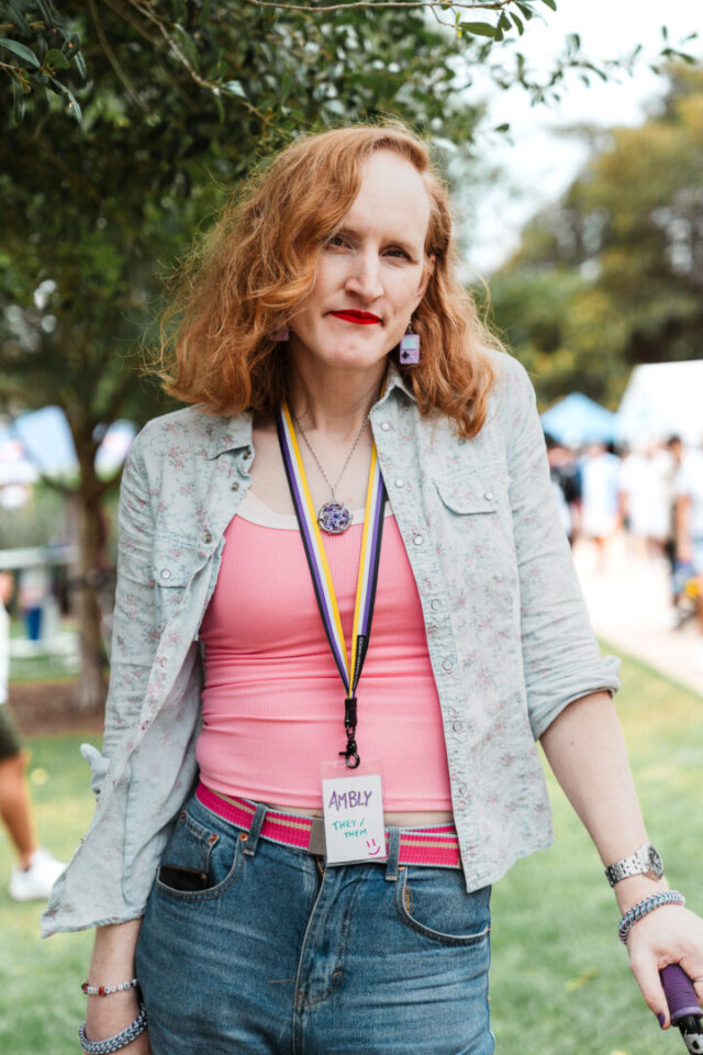 A person posing for a photo at Fair Day. They have medium length red hair and are wearing a pink singlet, open shirt and jeans.