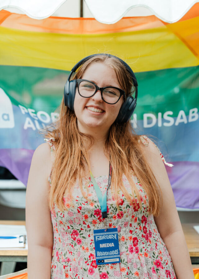 A person posing for a photo at Fair Day. They have long hair, wearing black reading glasses, headphones and a floral dress. A rainbow People with Disability Australia flag is visible in the background.