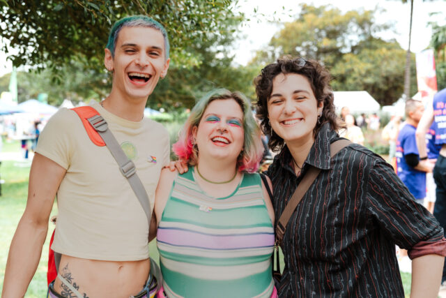 Three people with colourful hair, piercings and makeup, smiling and laughing at Fair Day. They have their arms around each other. There are trees and festival stalls in the background.