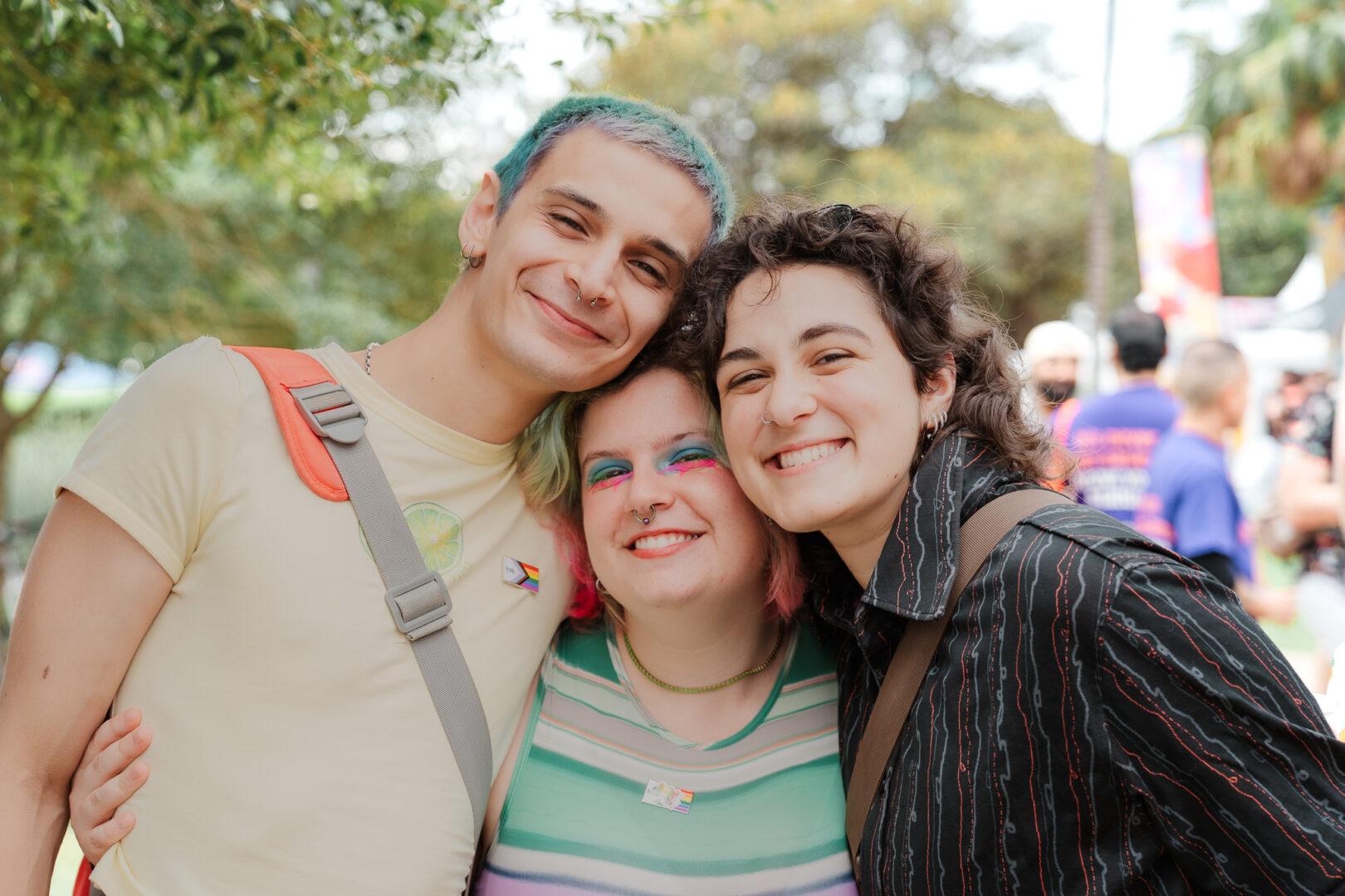 Three people with colourful hair, piercings and makeup, smiling at Fair Day. They have their arms around each other.