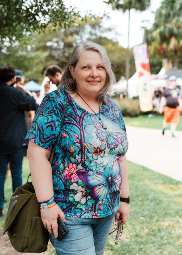 A middle aged person with shoulder length grey hair, wearing a blue butterfly print t-shirt and jeans posing for a photo at Fair Day.