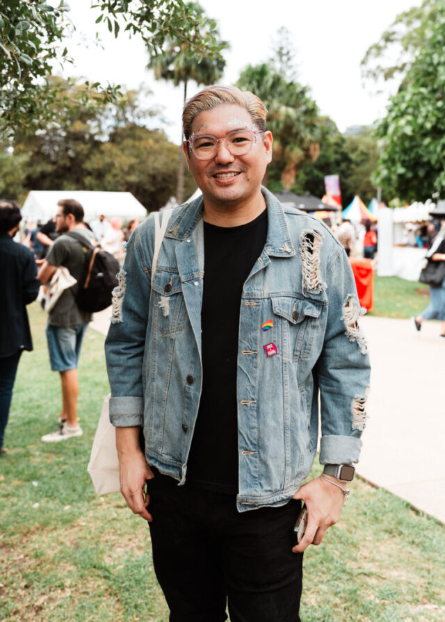 A person with short slicked backed blonde hair, smiling for a photo at Fair Day. They are wearing glasses and a distressed denim jacket with small rainbow pins.