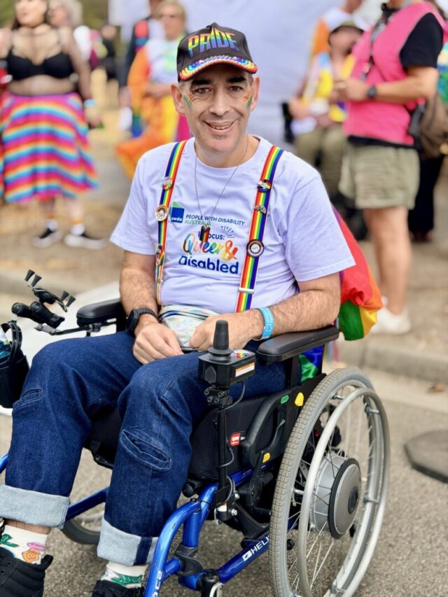 PWDA President Jeramy Hope at the Midsumma Pride March. Jeramy is sitting in a wheelchair and is wearing a PWDA pride t-shirt, jeans, rainbow suspenders.