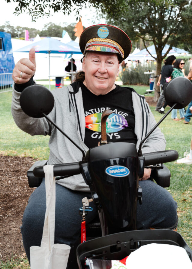 An older person at fair day. They are sitting on a mobility scooter and wearing a decorated cap and a black T-shirt with a rainbow ‘Mature Age Gays’ design. They are smiling at the camera and have their thumb up.