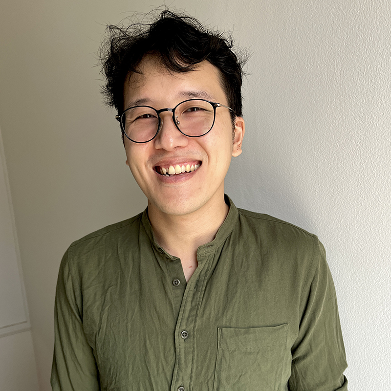 Smiling Asian man with round glasses in a green button-down shirt, standing against a light wall.