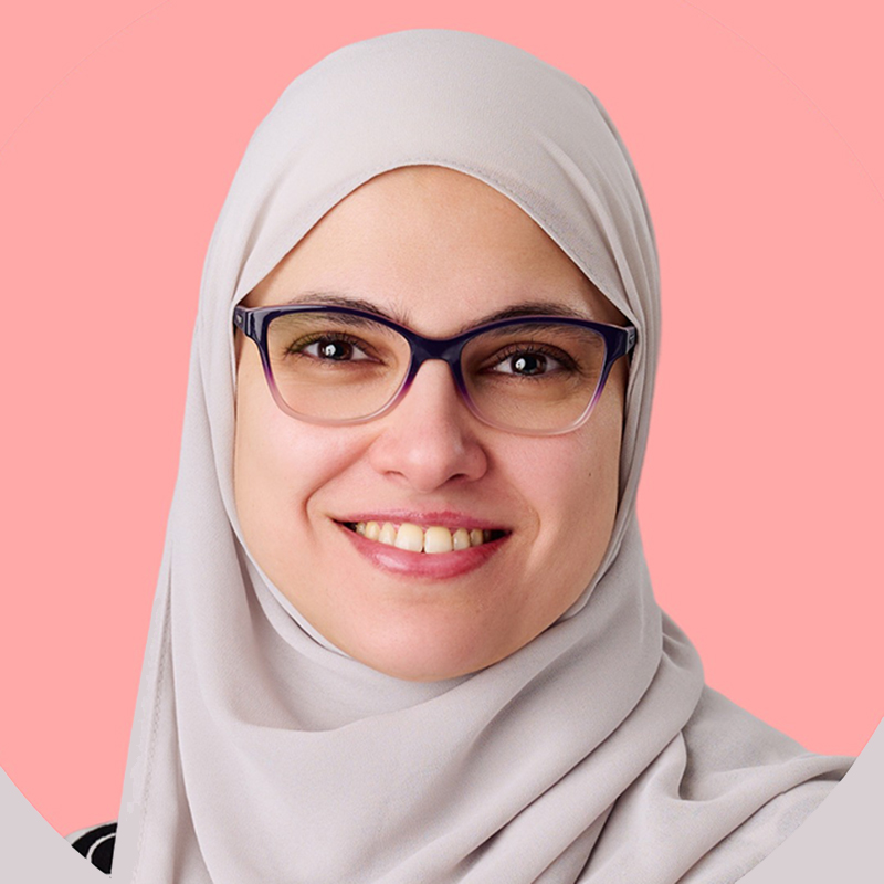Smiling woman wearing a light gray hijab and glasses against a pink background, looking at the camera.