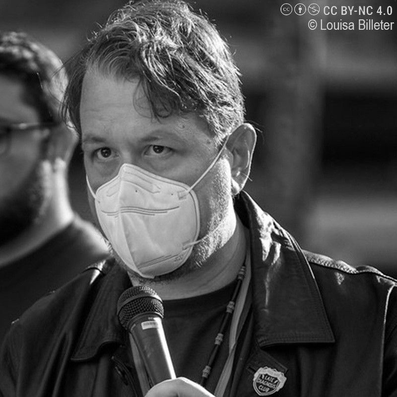 Man wearing a protective face mask speaks into a handheld microphone at an outdoor event, wearing a dark leather jacket and a beaded necklace.