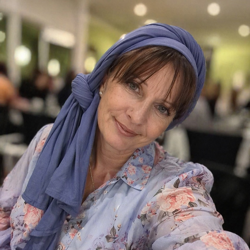 Smiling woman with a blue headscarf and floral blouse looking at the camera in a softly lit cafe setting.