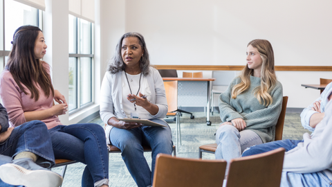 Three women sit in a circle in a bright meeting room, taking part in a group discussion. One woman gestures as she speaks while the others listen.